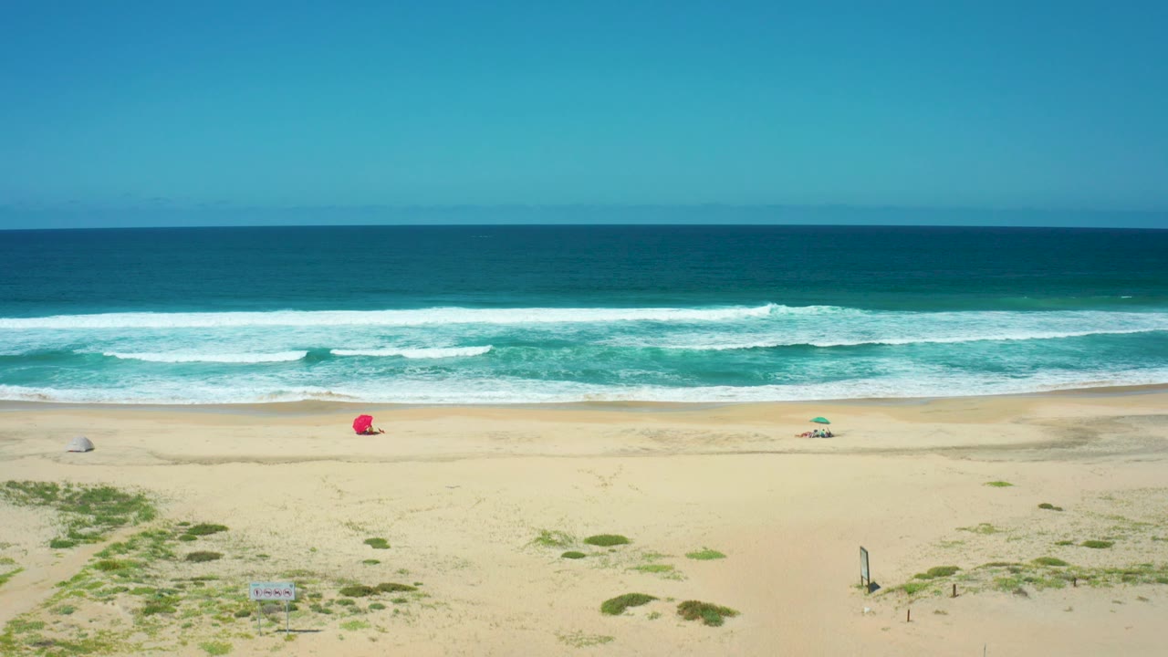 Aerial Cerritos Beach frame with rolling whitecaps, a wide sandy beach, low dune vegetation entering the lower foreground, a red object on the sand, and no visible people.