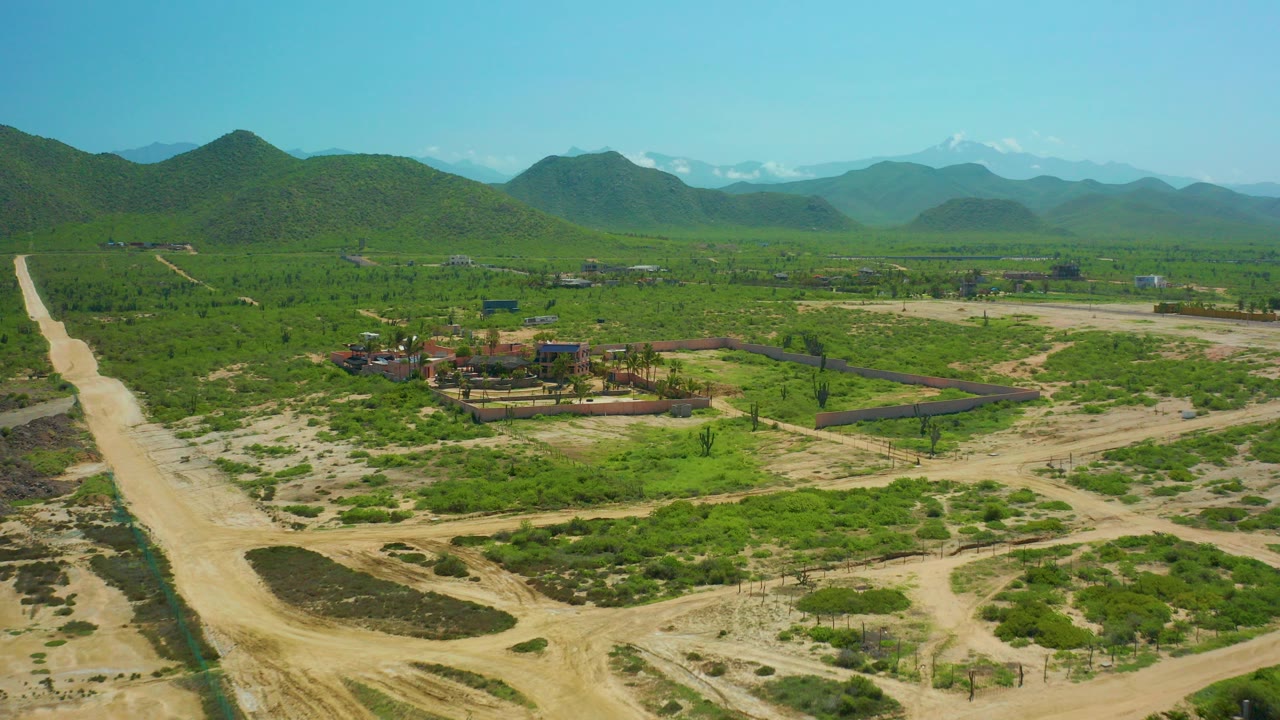 Aerial frame over rural Cerritos Beach, Baja California Sur with a wide dirt road at left, green vegetation, a small building cluster, distant mountains, clear sky, and no visible people.