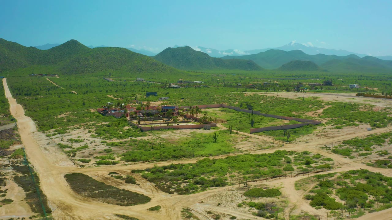 High aerial view of Cerritos Beach, Baja California Sur showing intersecting dirt tracks, green fields, a walled building area, distant peaks, clear sky, and no visible people.