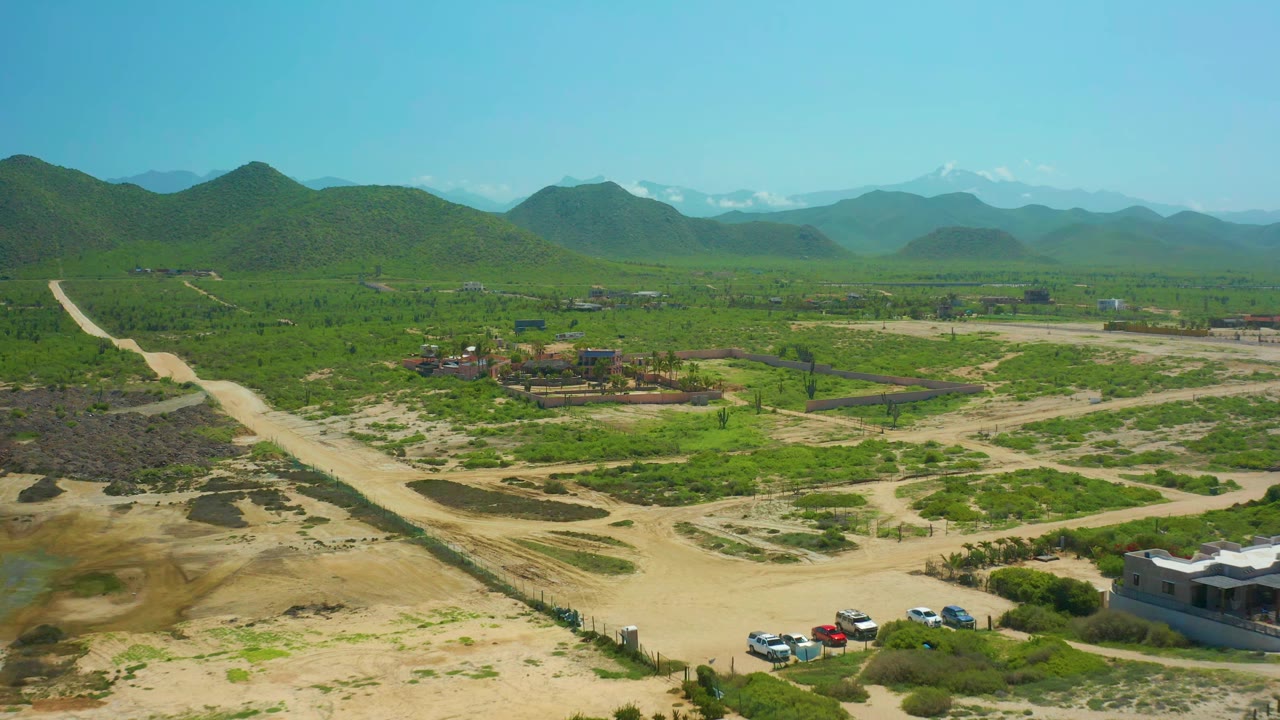 Aerial frame over Cerritos Beach rural terrain showing a pale dirt road, clustered vehicles near a building, green vegetation, rolling hills, far mountains, and no visible people.
