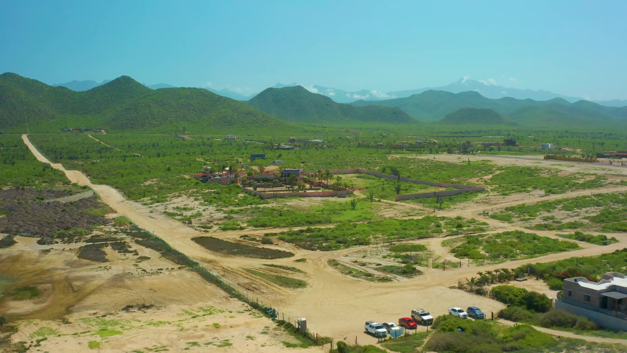Wide aerial view in Cerritos Beach, Baja California Sur, with a diagonal dirt road, scattered vehicles at the lower edge, open scrubland, green hills, distant mountains, and no visible people.