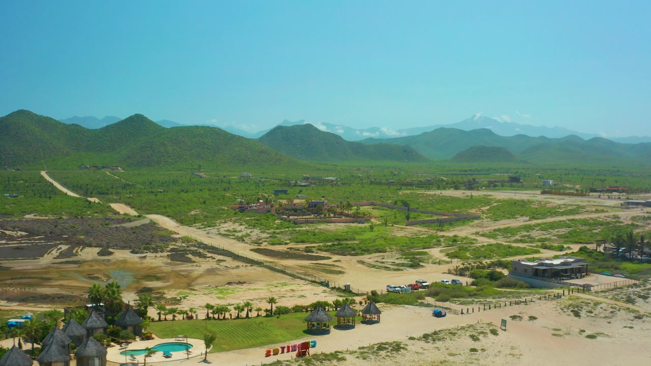 Aerial view over Cerritos Beach, Baja California Sur, showing green fields, sandy dirt roads, foreground huts and pool, distant mountains, and no visible people.