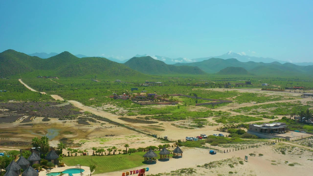 Wide drone view over Cerritos Beach rural land with sandy tracks crossing green fields, scattered buildings near the lower edge, mountains on the horizon, and no visible people.