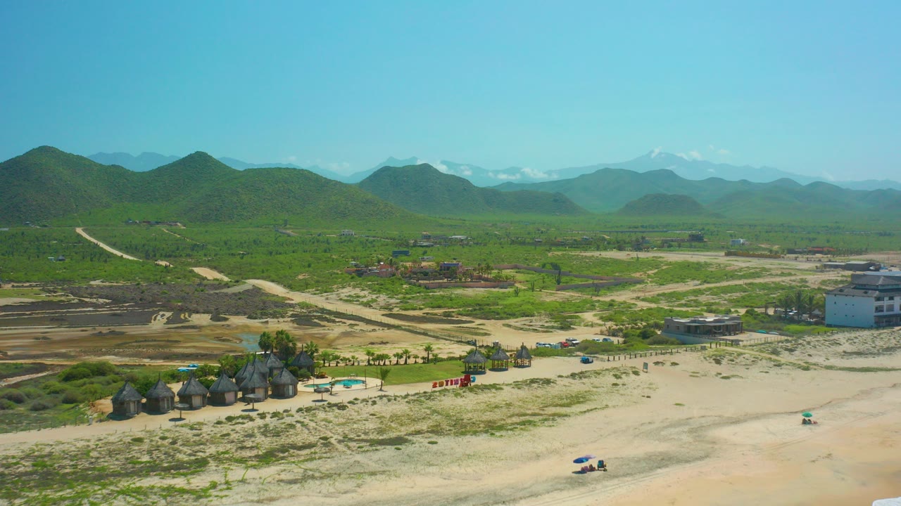 Aerial frame over Cerritos Beach BCS Mexico showing sandy foreground, thatched huts, dirt roads, green hills, distant snow-capped mountains, clear blue sky, and no visible people.