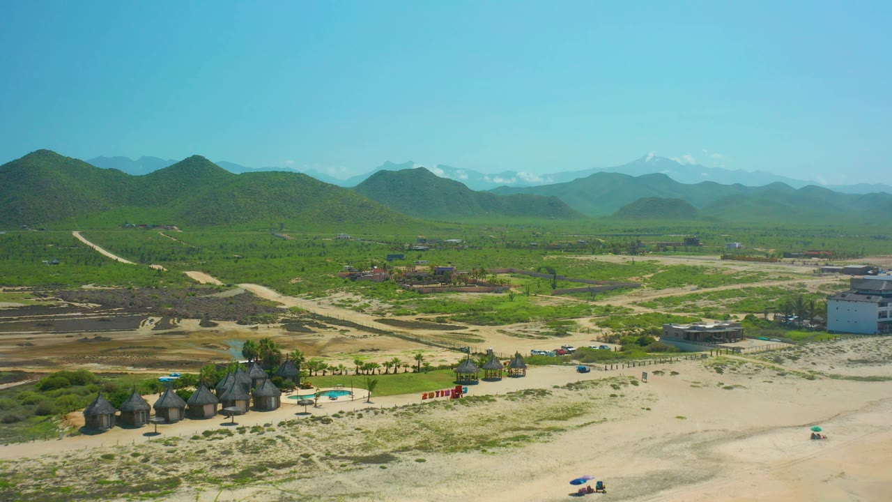 Cerritos Beach BCS Mexico aerial view showing huts near the lower edge, dirt roads crossing sandy and green ground, small buildings, vehicles, layered hills, distant snow-capped peaks, and no visible people.