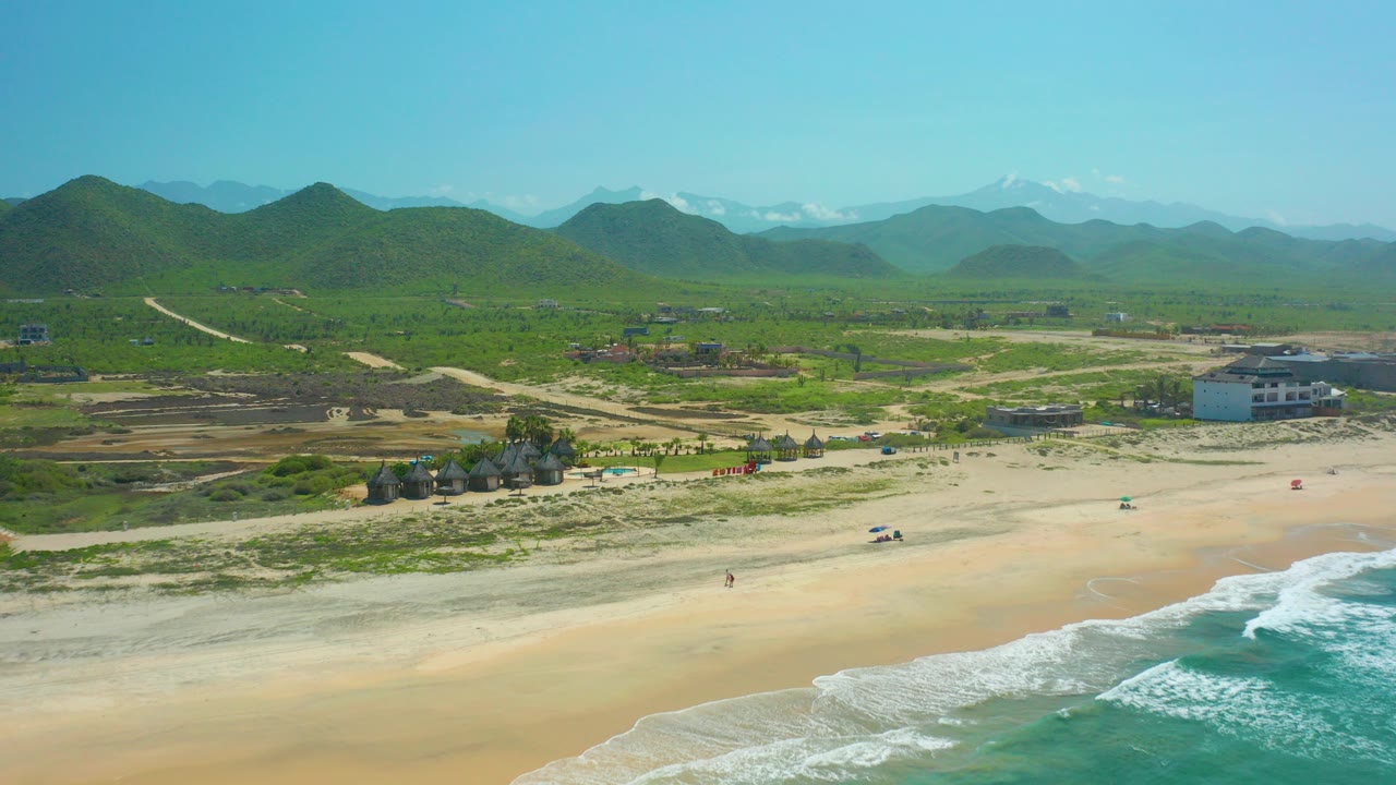 Aerial frame over Cerritos Beach in Baja California Sur showing pale sand, white surf at the lower right, beach structures, a coastal building, green hills, and distant mountains under clear sky.