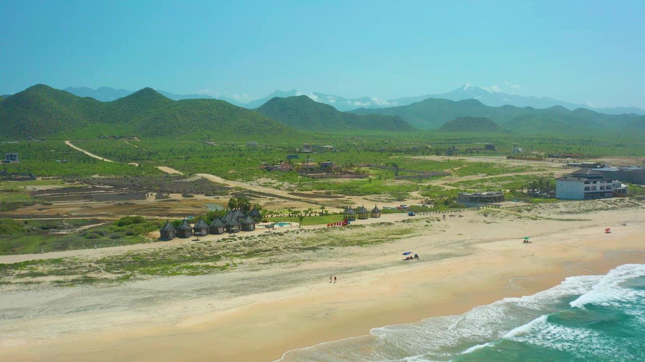 Aerial view of Cerritos Beach showing more open sand across the center, turquoise water at the lower right, low vegetation on dunes, scattered coastal buildings, and green hills beyond.