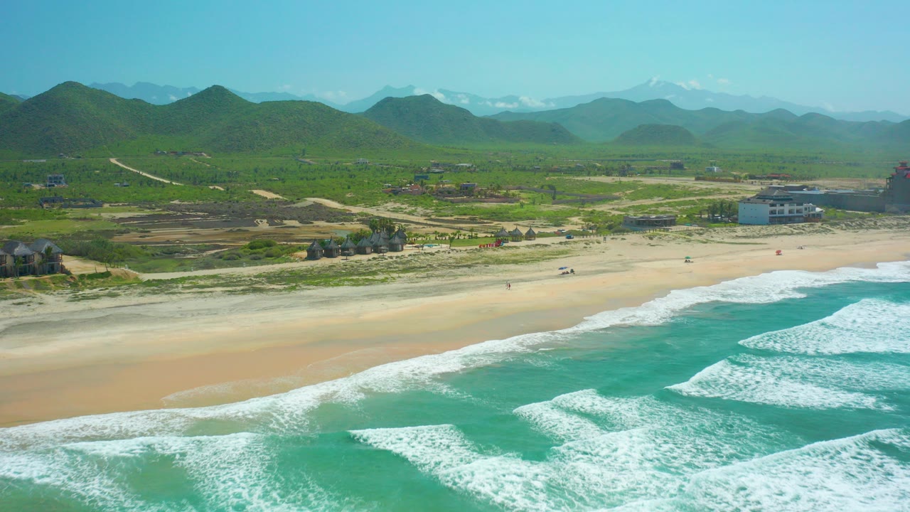 Wide aerial frame over Cerritos Beach, Baja California Sur, with turquoise surf and white foam in the foreground, pale sand in the middle distance, green hills and mountains beyond, and no visible people.