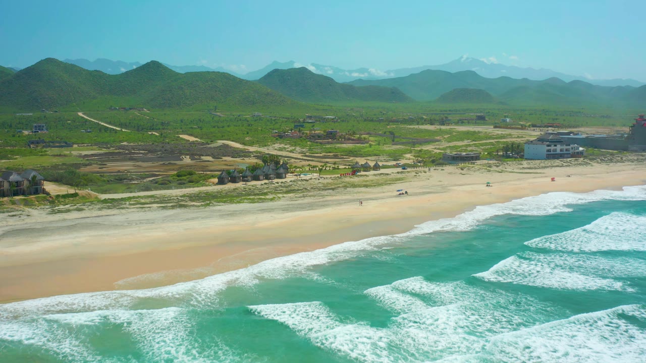 Turquoise surf breaks in parallel lines beside a broad sandy shore and low hills.