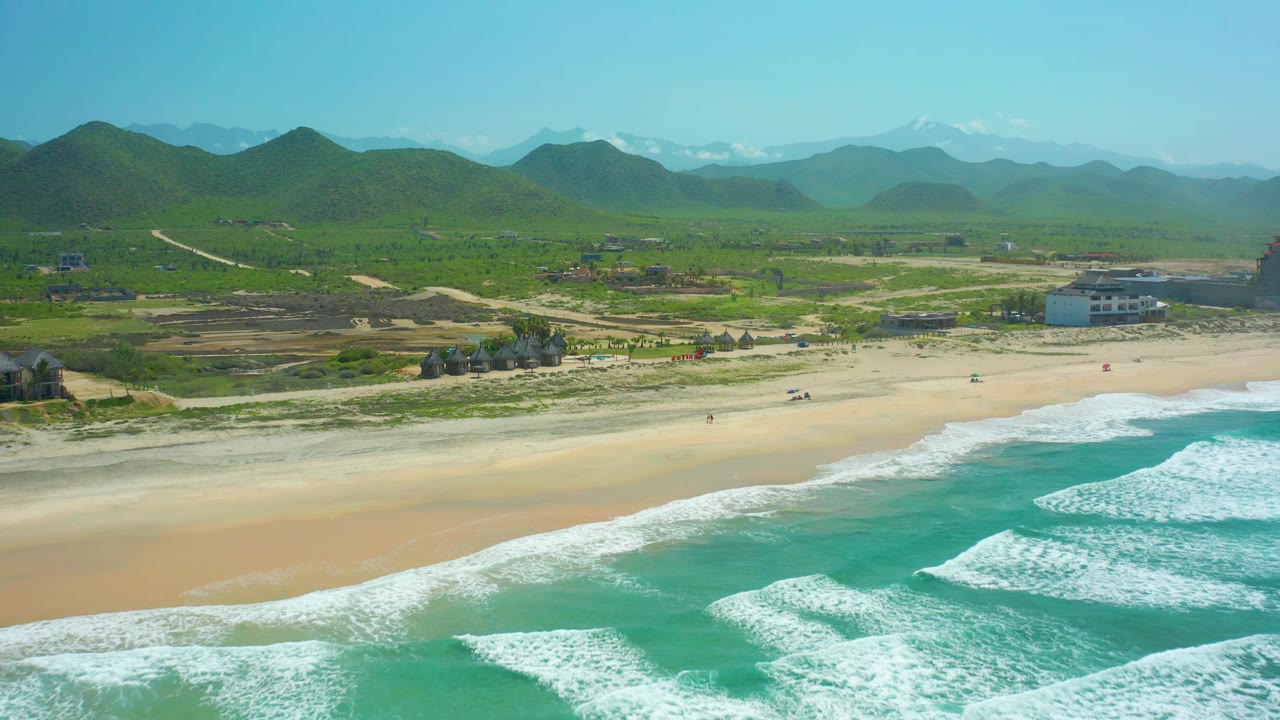Cerritos Beach appears from above with foam patterns closer to the lower right, a lightly marked sand beach across the center, low development near the shore, green hills inland, and no visible people.