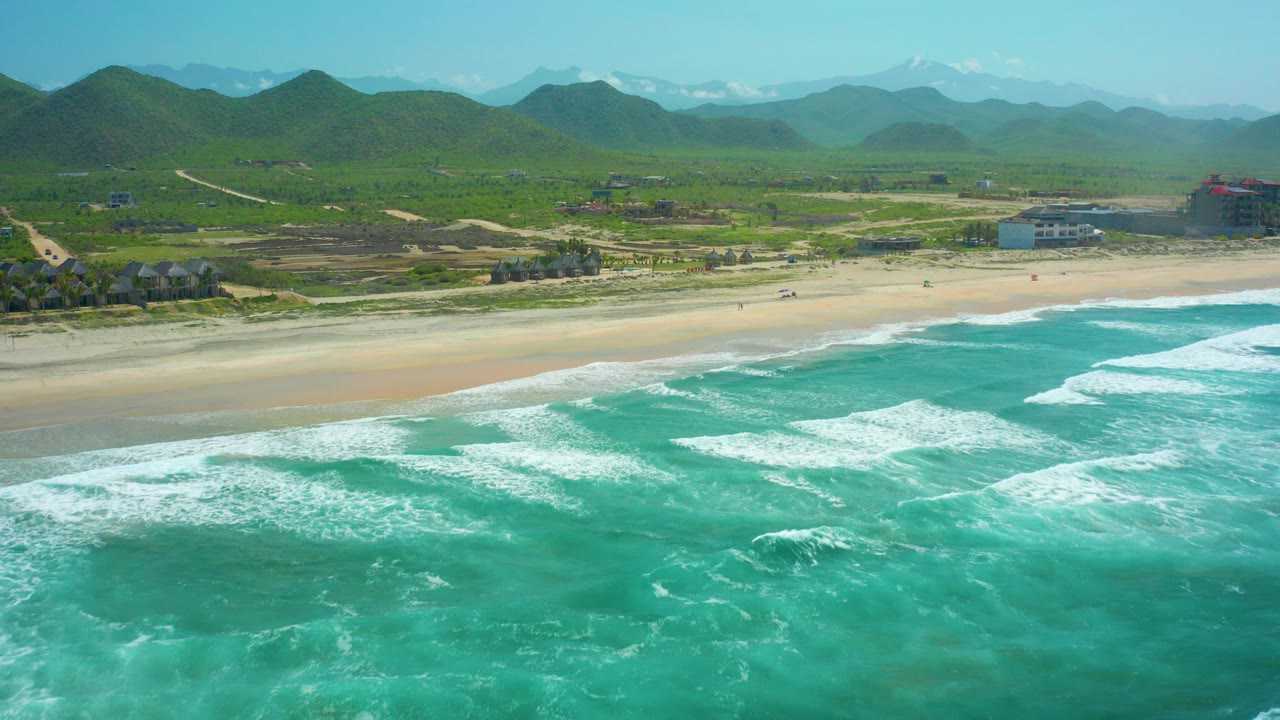 At Cerritos Beach in Baja California Sur, turquoise surf breaks across the foreground while a sandy beach, scattered buildings, green hills, and distant mountains fill the background with no visible people.