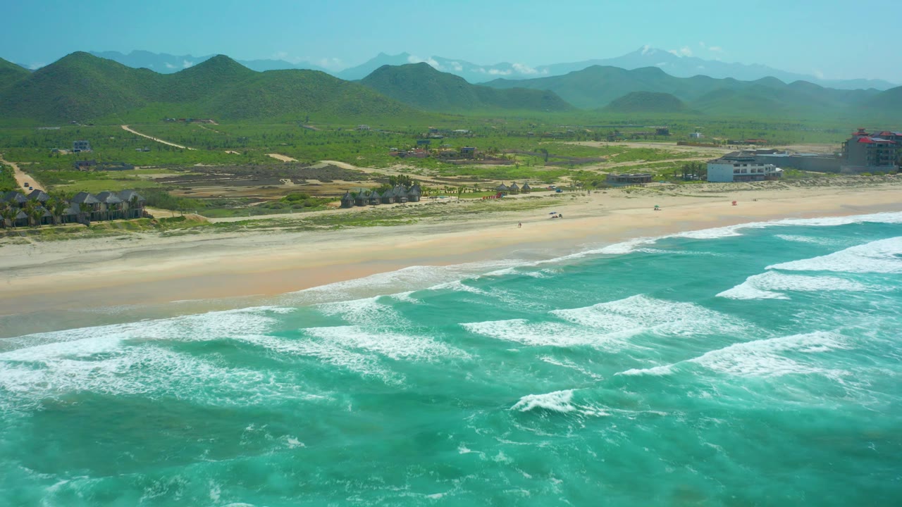 At Cerritos Beach, angled white surf lines lead toward a quiet sandy shoreline, with a small cluster of buildings, green vegetation, and layered hills visible inland, with no visible people.