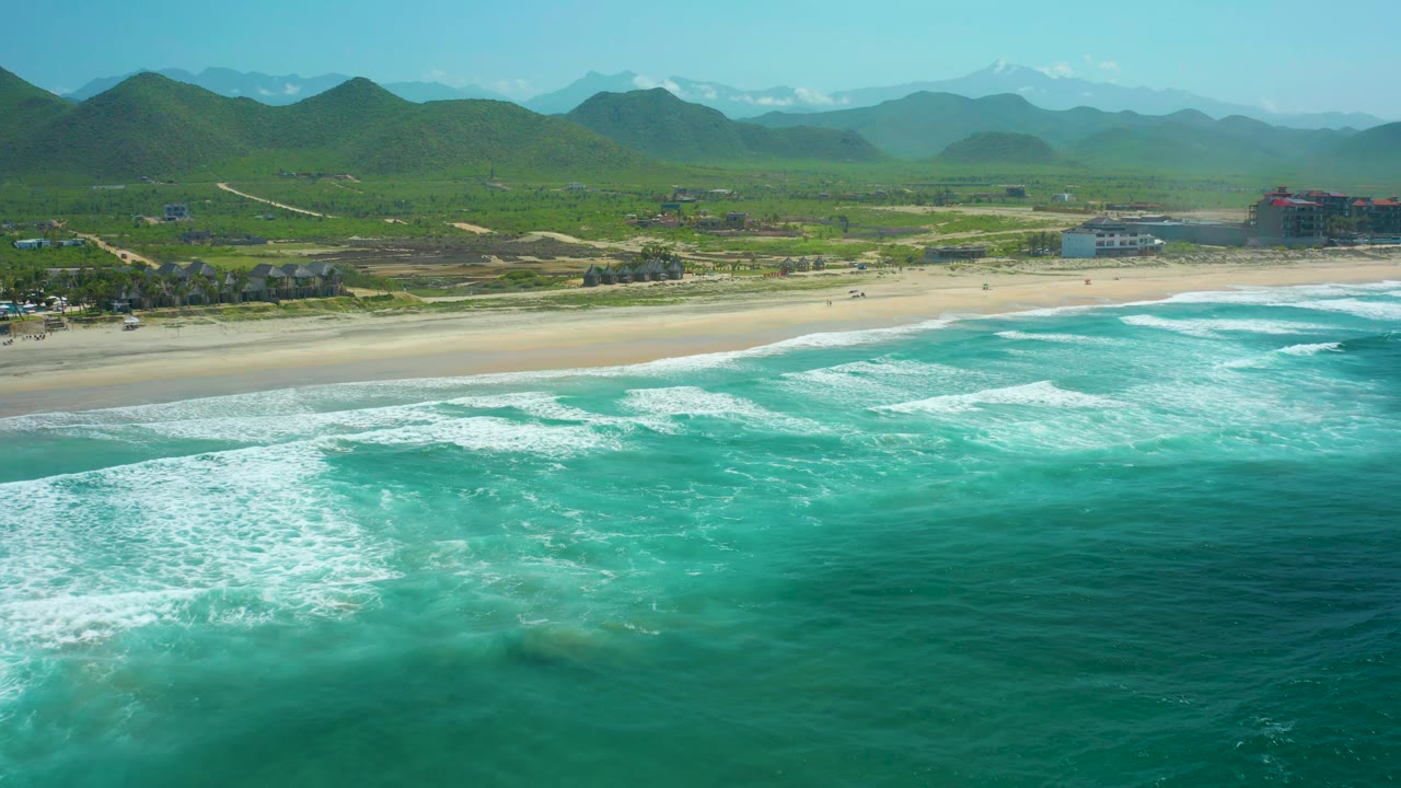 Wide aerial coastline with white surf bands, sand, green hills, and buildings on the right.
