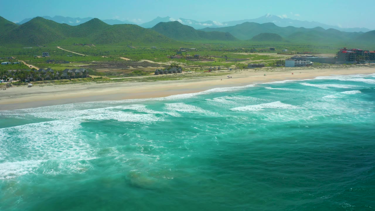 Aerial view at Cerritos Beach with rolling turquoise waves closer to shore, open sand across the middle of frame, layered green mountain ridges, coastal buildings to the far right, and no visible people.