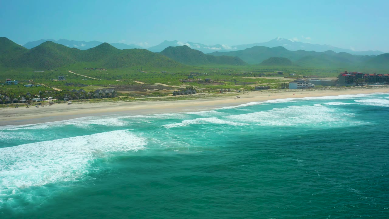 At 0:01, the Cerritos Beach aerial view shows a broader band of white foam crossing the turquoise ocean, with pale sand, small distant buildings, green hills, and no visible people.