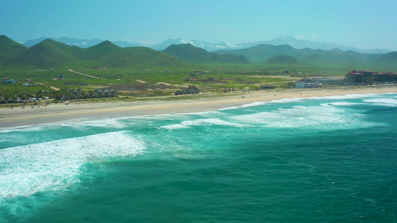 At 0:02, Cerritos Beach remains framed from above with foamy surf rolling toward the sand, green hills behind the beach, distant low buildings at the right, and no visible people.