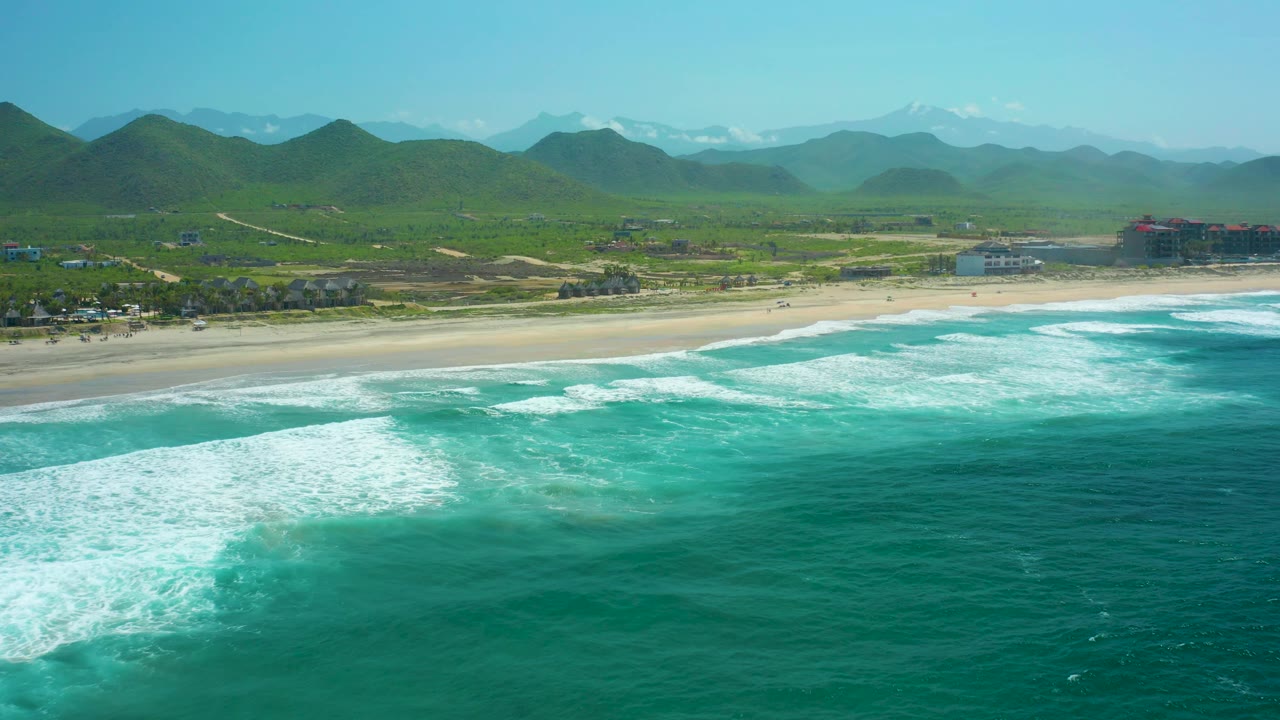At 0:03, the aerial frame shows white foam bands nearer the lower left foreground, the sandy Cerritos Beach shoreline through the middle, green hills beyond, and no visible people.