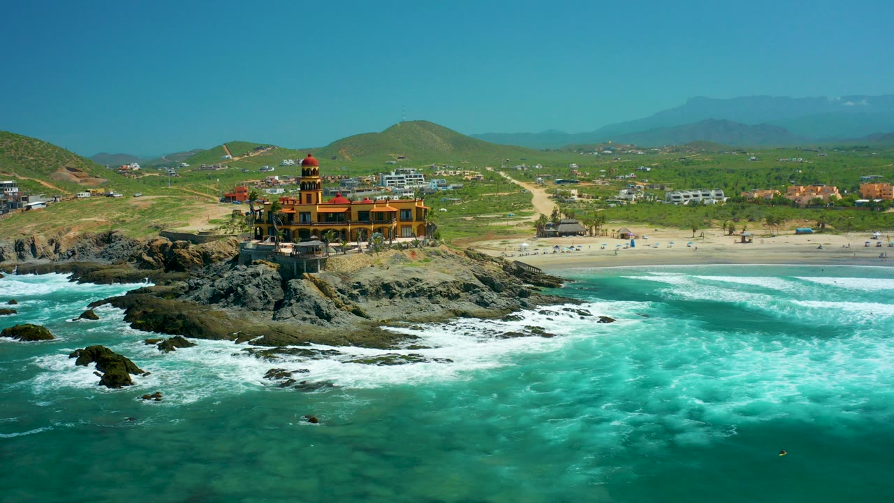 Aerial frame at Cerritos Beach, Baja California Sur, showing turquoise surf around dark rocks, an orange lighthouse-like building on the outcrop, sandy beach and green hills behind it, with no visible people.
