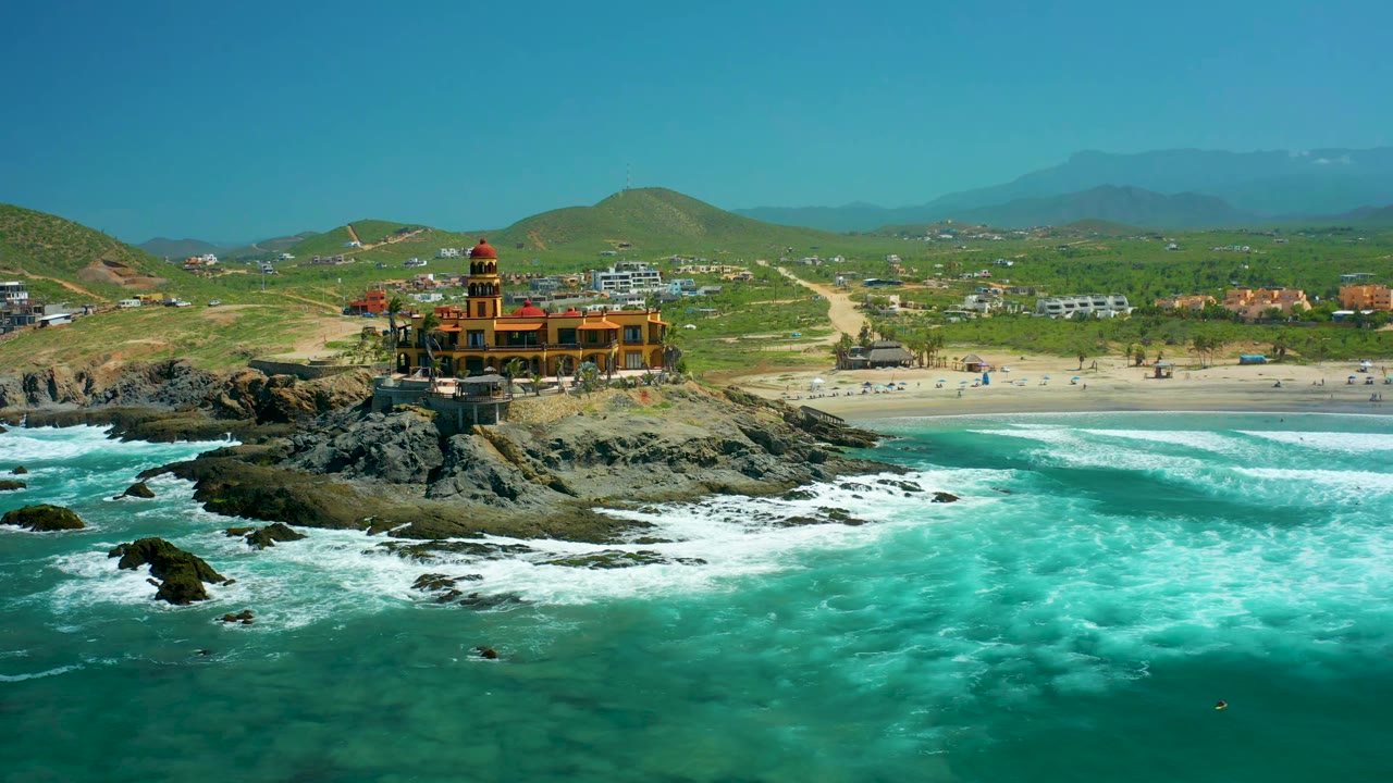 At 0:01, the Cerritos Beach aerial view holds the lighthouse-like building near the rocky point while a broad band of white foam spreads across the turquoise water, with no visible people.