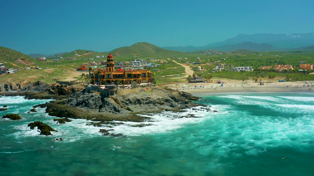 At 0:02, waves break around the rocky outcrop below the orange lighthouse-like structure, with the Cerritos Beach sand, low buildings, green hills, and hazy mountains visible beyond, and no visible people.