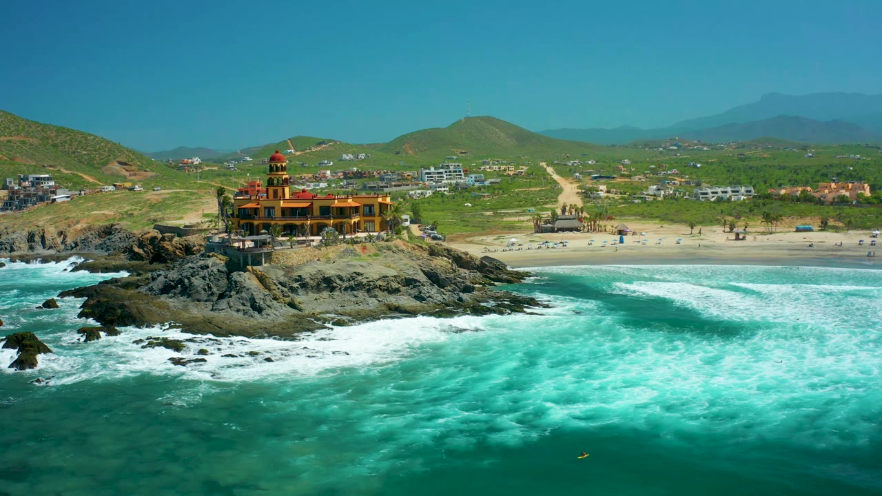 At 0:01, the Cerritos Beach lighthouse remains centered on dark rocks as a wider band of white foam spreads across the turquoise foreground, with no visible people.