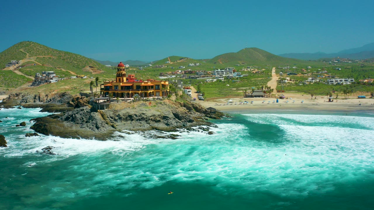 At 0:00 in Cerritos Beach, Baja California Sur, a yellow-orange building sits on a rocky outcrop above turquoise surf, with sandy beach, green hills, clear sky, and no visible people.