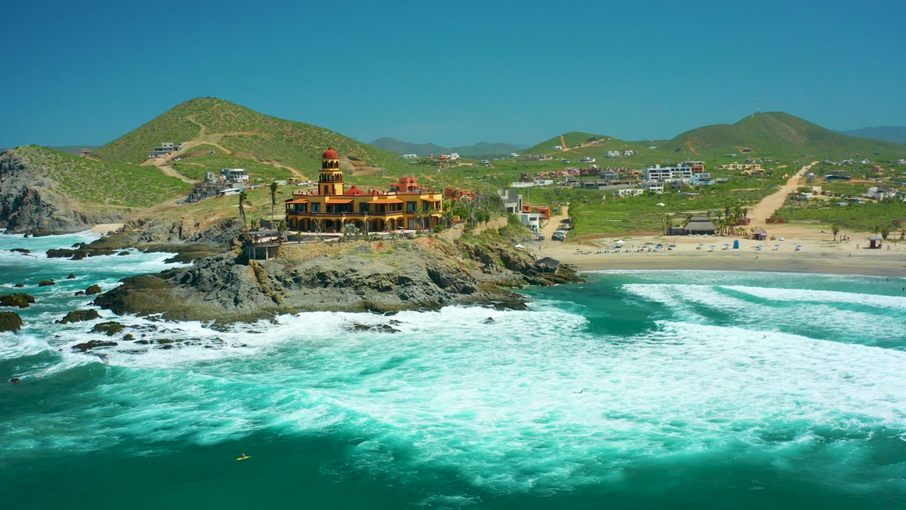 At 0:00, Cerritos Beach appears from above with white surf in the foreground, a temple-like yellow and red building on dark rocks, sandy beach beyond, green hills, and tiny visible people on the beach.