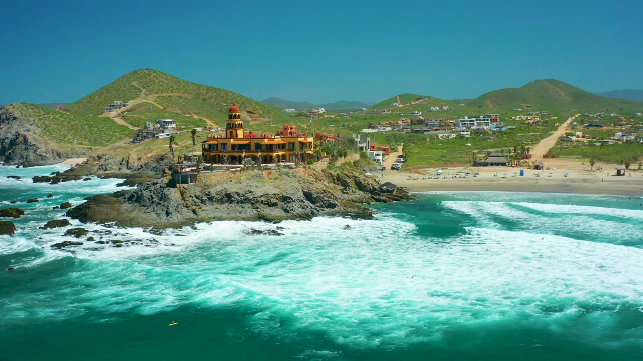 Drone view of waves spreading across turquoise water below a rocky temple-like coastal building.