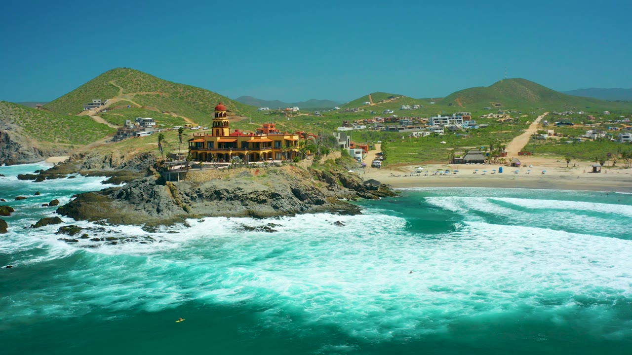 At 0:02, the Cerritos Beach coastline remains in a wide aerial frame as surf wraps around the rocks, the red-roofed temple-like building sits near center, and scattered beachgoers appear small on the sand.