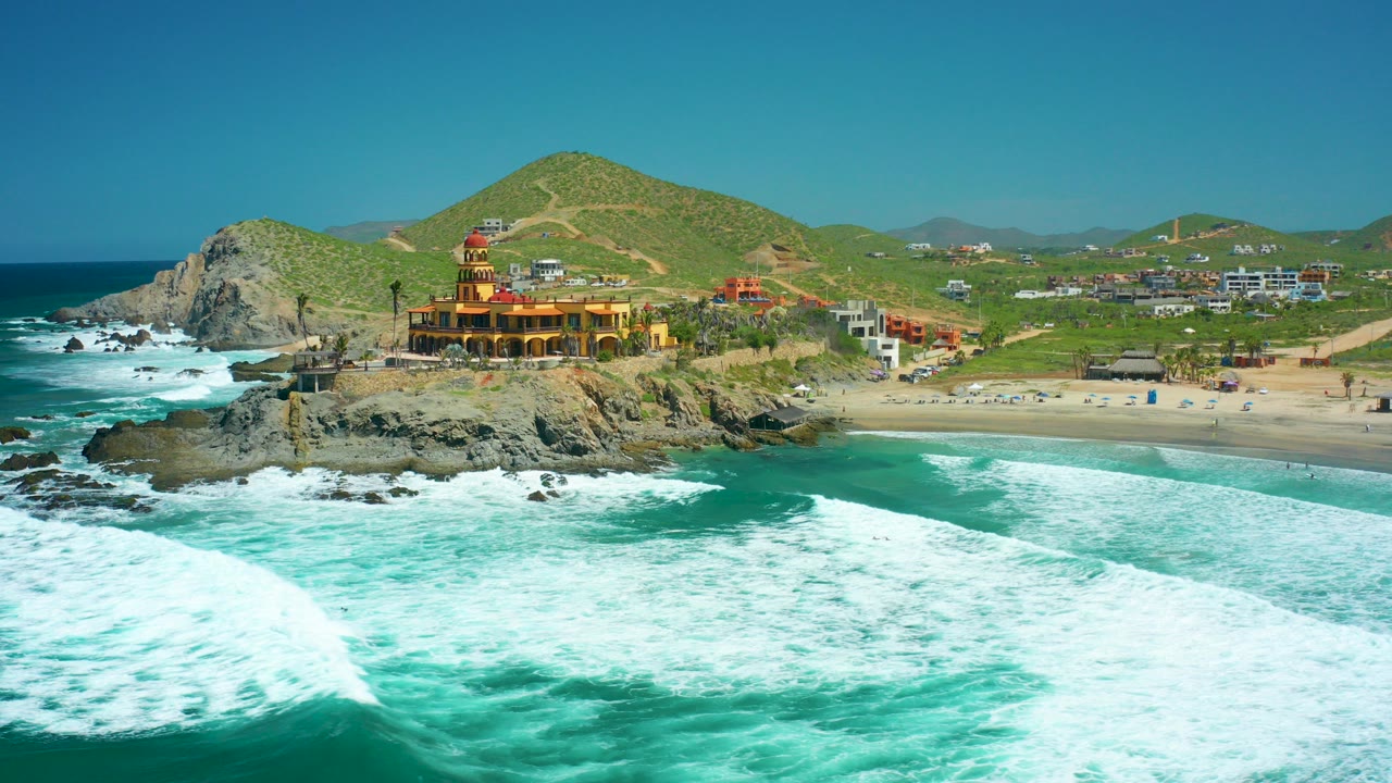 At Cerritos Beach in Baja California Sur, turquoise waves break across the foreground while a yellow coastal building sits on dark rocks below green hills, with no visible people.