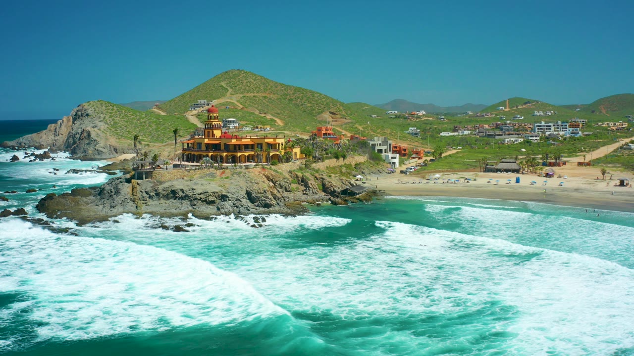 Aerial coastal view with foamy waves, rocks, a beach building, and green hills.