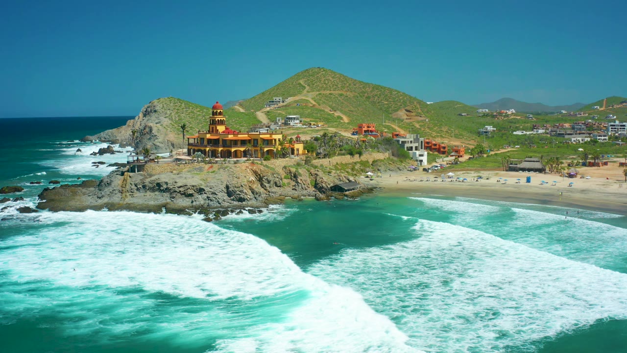 At 0:01, a broad band of white foam advances toward the beach at Cerritos Beach while the rocky outcrop, coastal structure, hillside roads, scattered buildings, and no visible people remain in view.