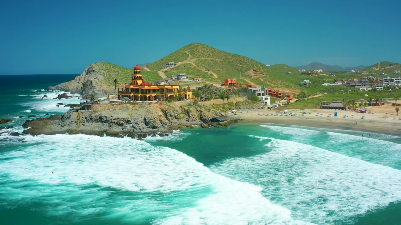 At 0:02, the wave line stretches diagonally across the foreground at Cerritos Beach, with the rocky headland, lighthouse-like structure, beach parking area or small structures, green hills, and no visible people behind it.