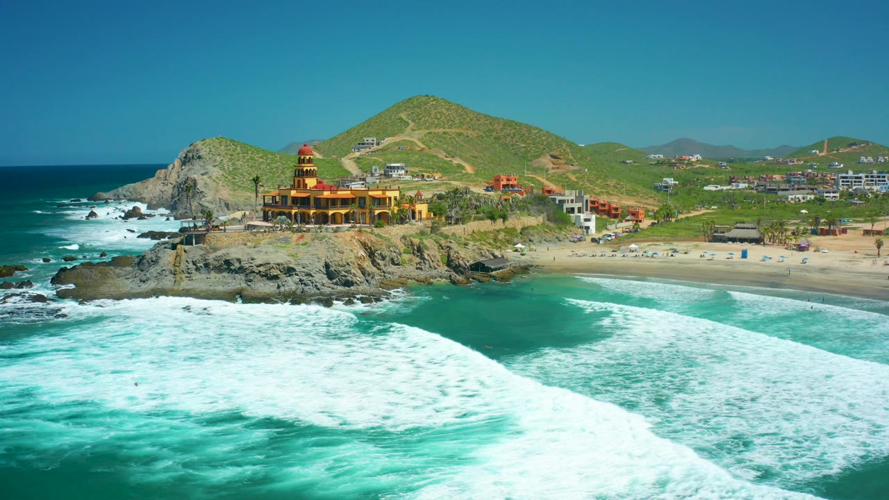Rocky coastal point with surf, beach, hills, and a coastal building.