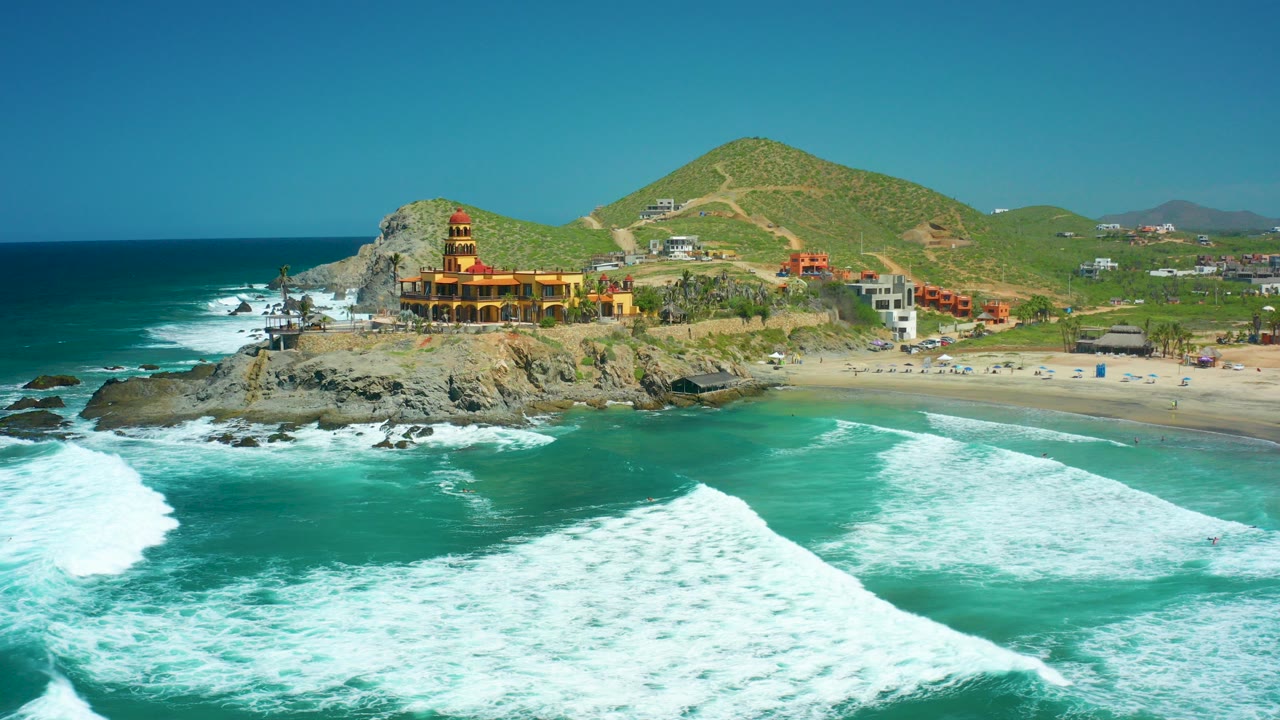 At Cerritos Beach in Baja California Sur, turquoise waves break across the foreground while a rocky point with a yellow and red lighthouse-like building sits below green hills, with no visible people.