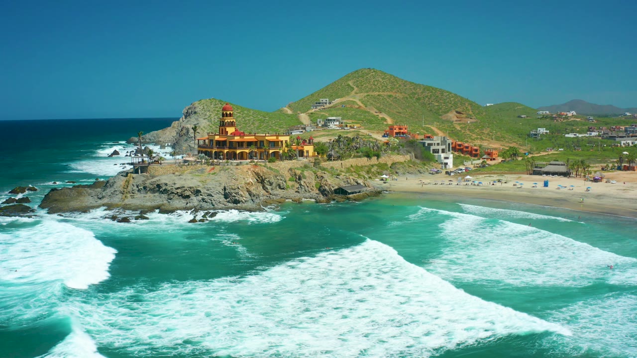 At 0:02, the aerial view holds the lighthouse-like building on the rocky outcrop as white surf bands sweep across the turquoise water toward the Cerritos Beach shoreline, with no visible people.