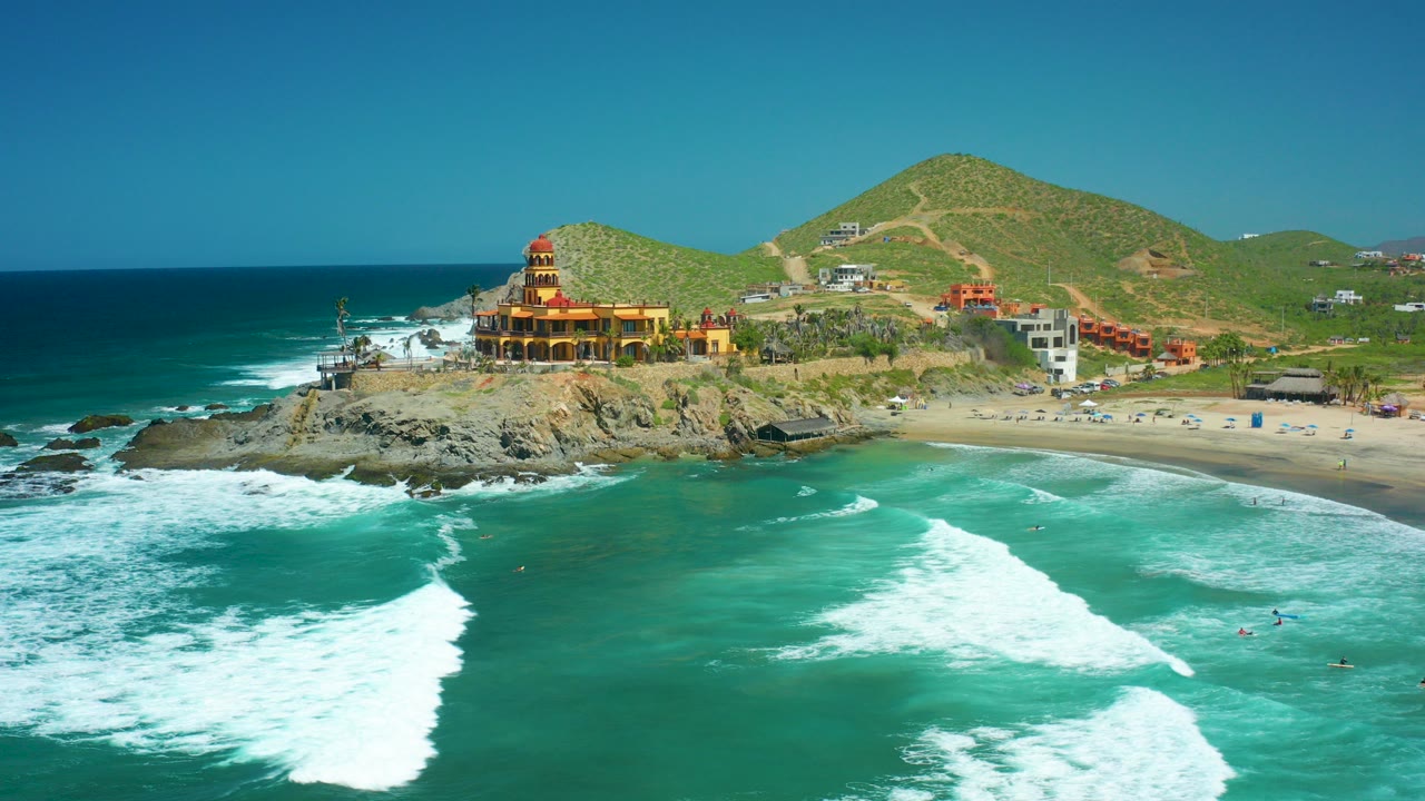 Opening aerial frame at Cerritos Beach, Baja California Sur, with turquoise surf breaking across the foreground, a yellow coastal building on rocks, green hills behind, and no visible people.