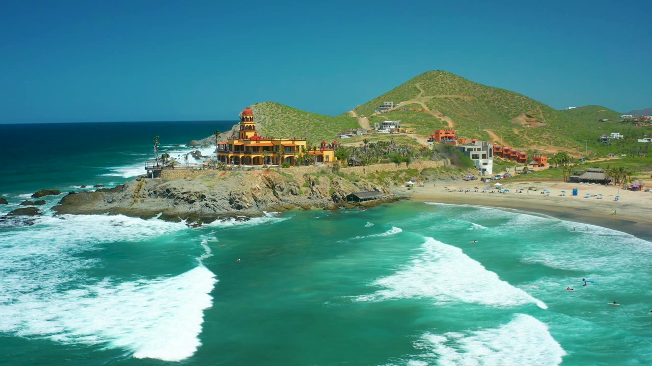 Aerial frame at Cerritos Beach showing a broad fan of white foam moving toward the beach, the rocky point with a coastal building, hillside paths, and no visible people.