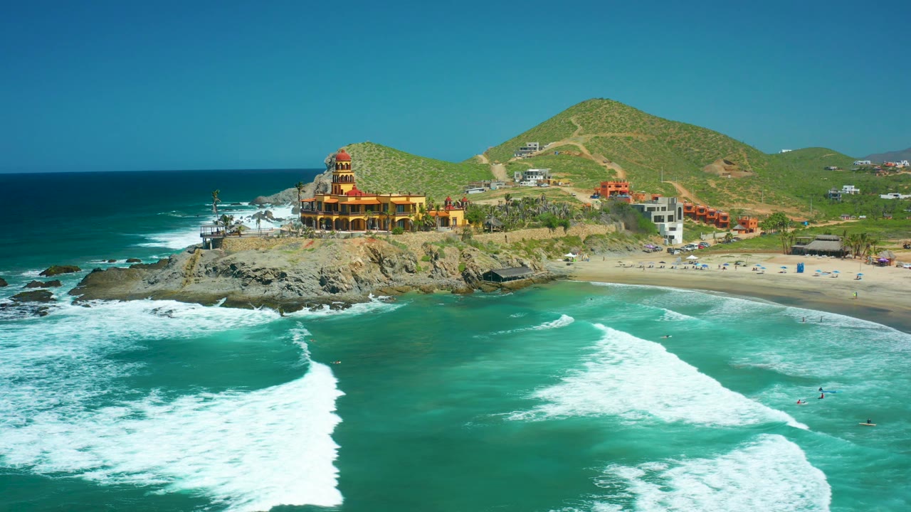 Cerritos Beach aerial frame with the surf line spread wider across the foreground, dark rocks beneath the coastal building, a sandy beach to the right, and no visible people.
