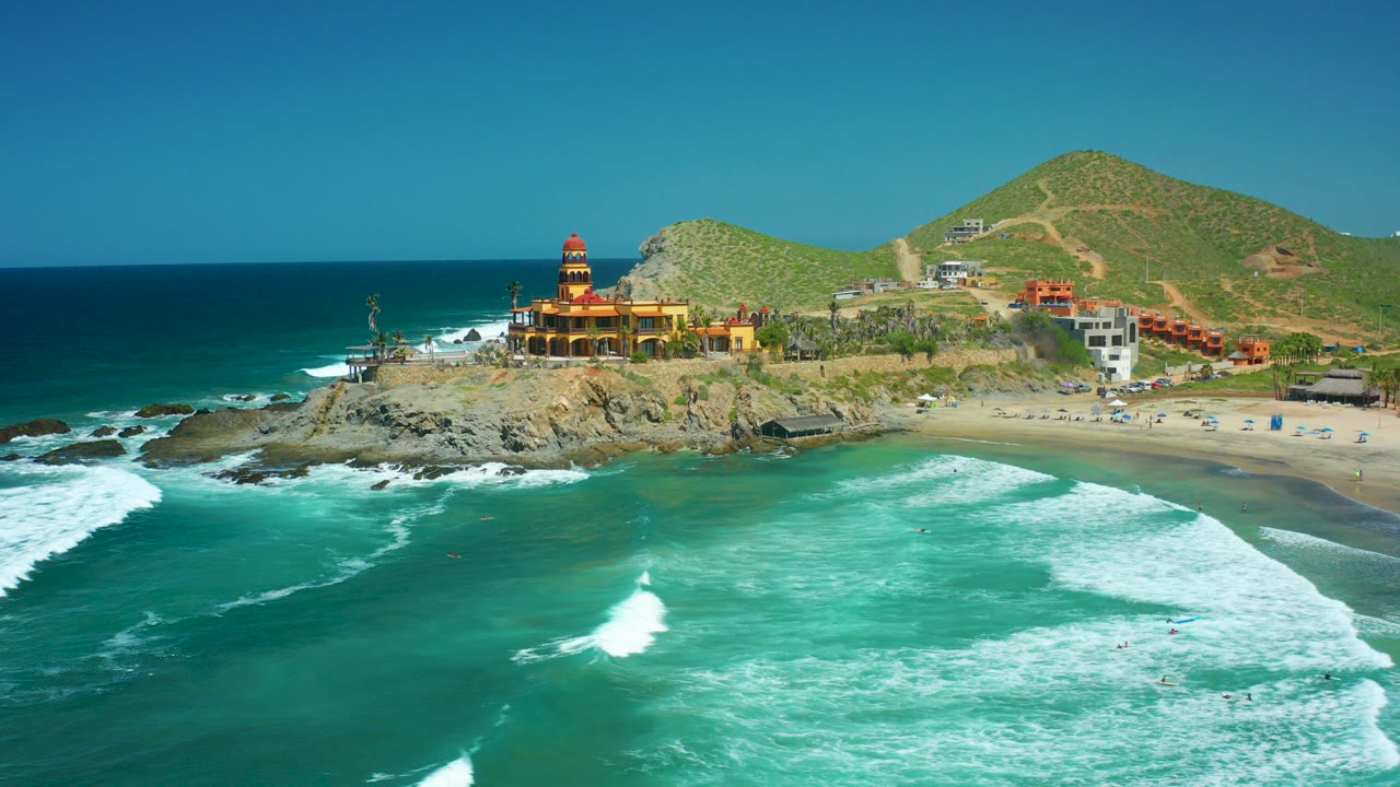Aerial frame of Cerritos Beach, Baja California Sur, with turquoise waves in the foreground, a lighthouse on a rocky point, beachgoers as small figures, and green hills behind.