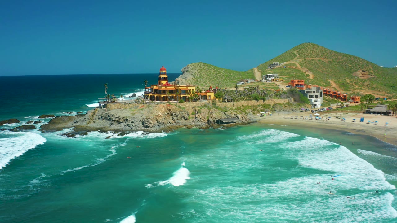 Wide aerial of waves rolling toward a sandy beach beside a lighthouse on rocks.
