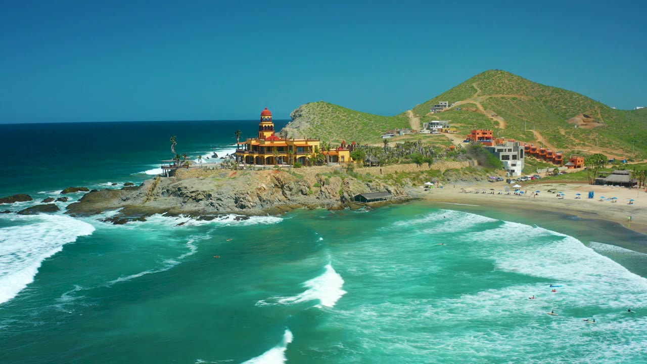 Cerritos Beach aerial frame with a large white breaker in the lower water, the lighthouse on the rocky coast, and green hillside roads and buildings in the background.