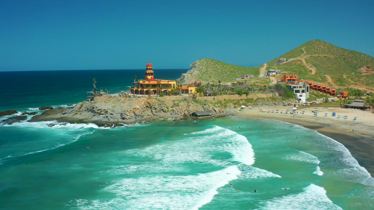 Aerial view of Cerritos Beach, Baja California Sur with a lighthouse on a rocky point, turquoise water, white surf, green hills, coastal buildings, and no visible people.