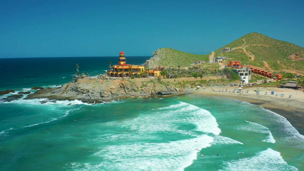 Aerial frame over Cerritos Beach showing the lighthouse on the rocky point, a larger foam pattern across the foreground water, beach umbrellas or small beach objects on sand, hillside buildings, and no visible people.