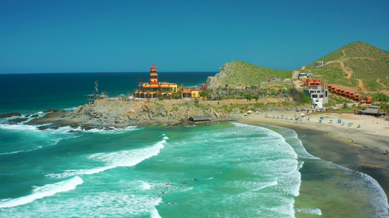 Wide aerial coastline with waves breaking toward sand and a lighthouse on rocks.