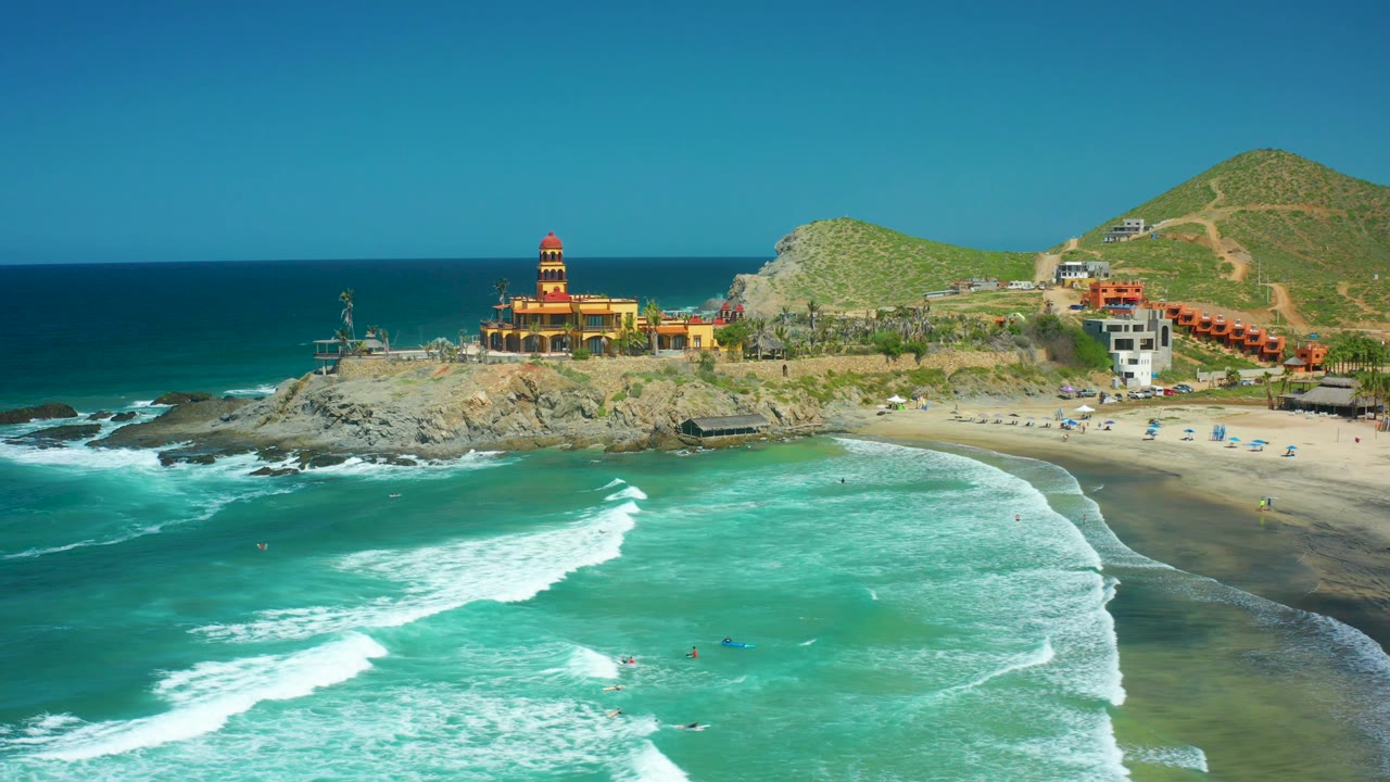 At 0:02, the Cerritos Beach shoreline shows thicker white foam across the lower frame, a darker blue ocean beyond the rocks, the lighthouse on the point, and no visible people.