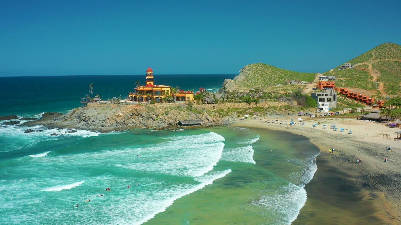 Aerial view over Cerritos Beach, Baja California Sur, with turquoise surf rolling toward sand, small swimmers offshore, a rocky point, a towered building, and green hills behind.