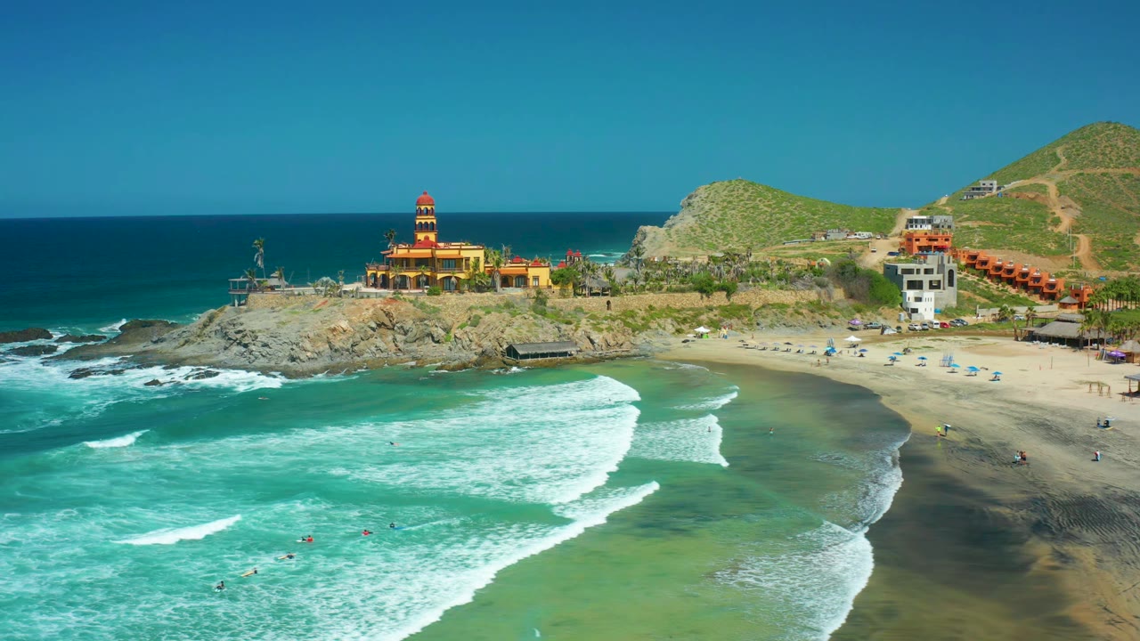 At 0:01 over Cerritos Beach, white surf lines push farther across the turquoise water while swimmers remain small in the foreground and the towered cliff building stays centered beyond the beach.