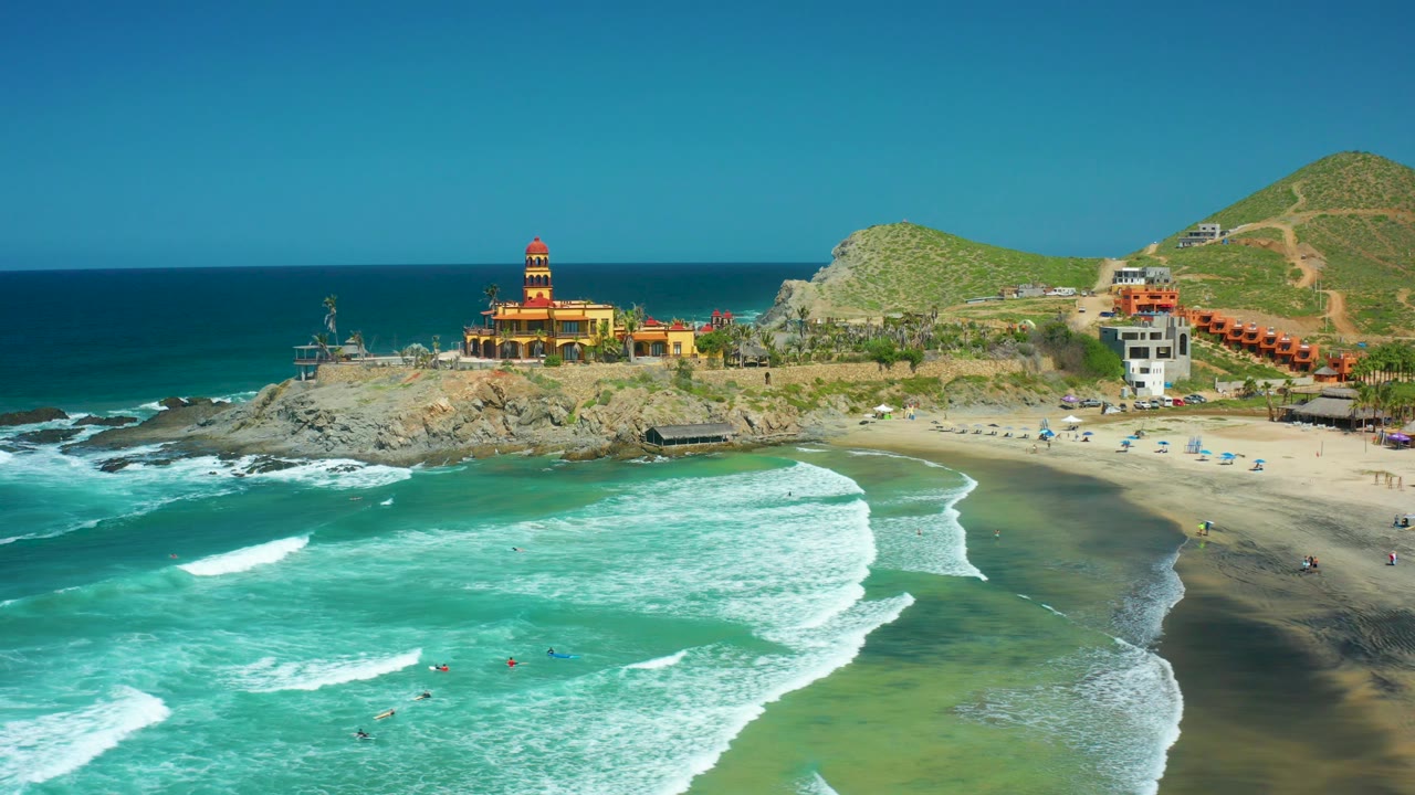 Aerial coastline view with foamy waves, swimmers, sandy beach, rocks, and hill buildings.