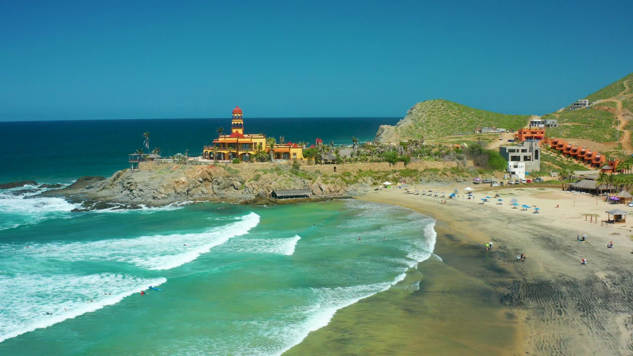 Aerial view of Cerritos Beach in Baja California Sur with turquoise surf breaking across the foreground, scattered beachgoers on the sand, and a lighthouse-style building on the rocky point.